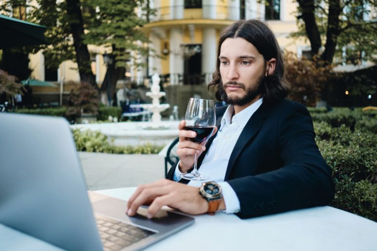 Handsome latin businessman with glass of wine thoughtfully working on laptop in restaurant outdoor